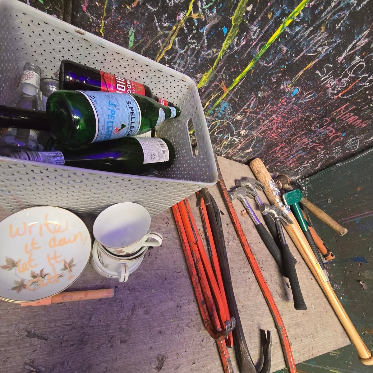 Art supplies and materials including paint bottles, brushes, ceramic dishes, and sketch paper arranged on a work surface with colorful painted backdrop