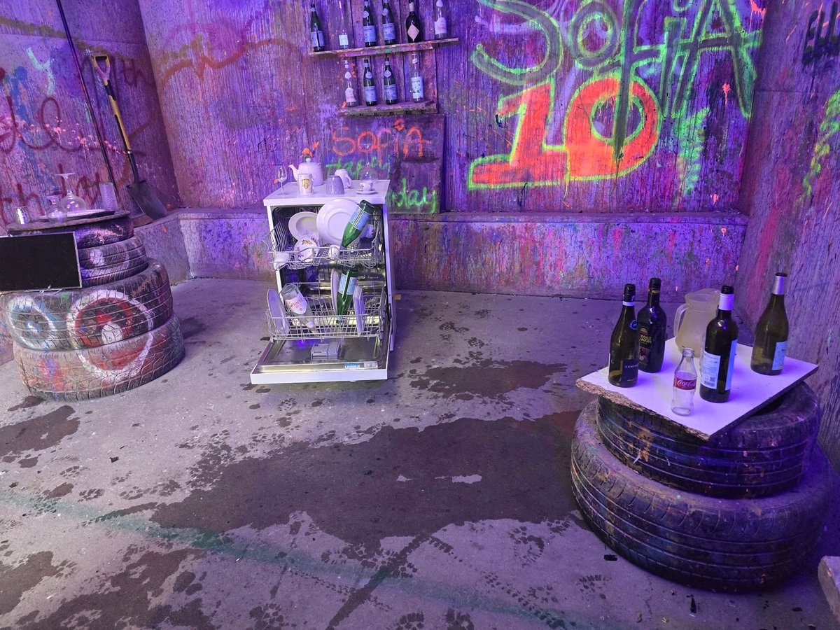 Purple-lit indoor space with neon signs, display cases, wine bottles on a tire platform, and vintage electronics in an abandoned-looking setting